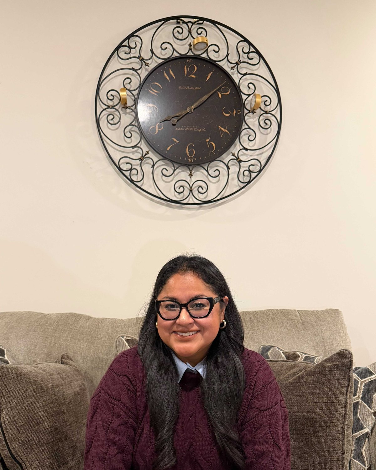 Flor Villagran smiling, seated on a couch beneath a large wrought-iron wall clock