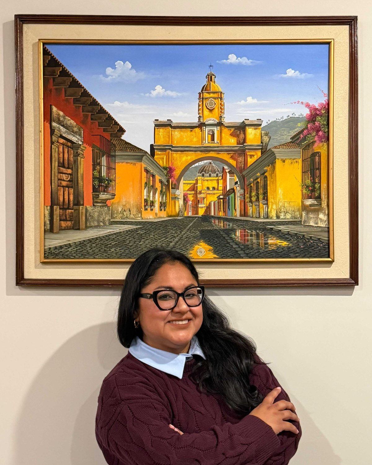 Flor Villagran smiling with her arms crossed in front of a painting of a yellow arch over a cobblestone street